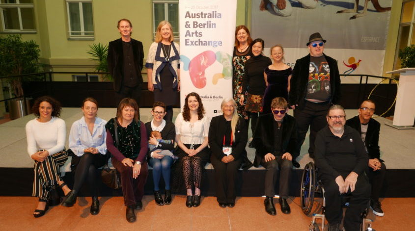 A group of people pose on stage, one row standing and one row sitting. In the middle is the Arts Exchange banner.
