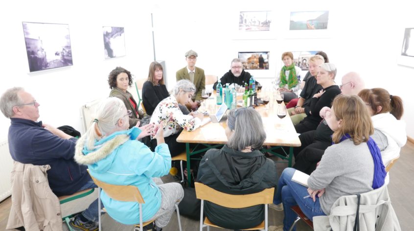 A group of fifteen Australian and Berlin-based artists sit around a table, deep in discussion. The walls display photographs which are part of an exhibition by artist Micha Winkler.