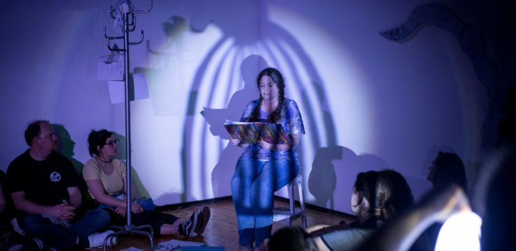 A woman sits on a chair in the corner of a room, reading aloud from a book. Several people sit on the floor, listening. A shadow of a large cage is cast over the reader.