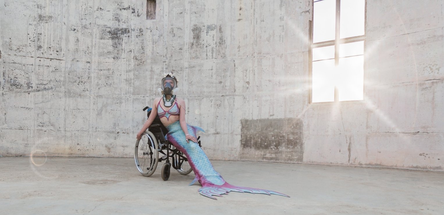 Image shows a woman dressed as a mermaid sitting in a wheelchair in an abandoned urban space. She wears a gas mask and neck brace. The sun flares through the ruins of a window