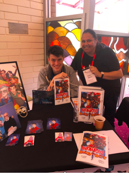 A boy in his late teens sits at a table filled with comic books and pictures of him meeting Stan Lee. Next to him stands a woman wearing black with long dark hair tied back. He holds two books in his hands, one from Be My Koorda.