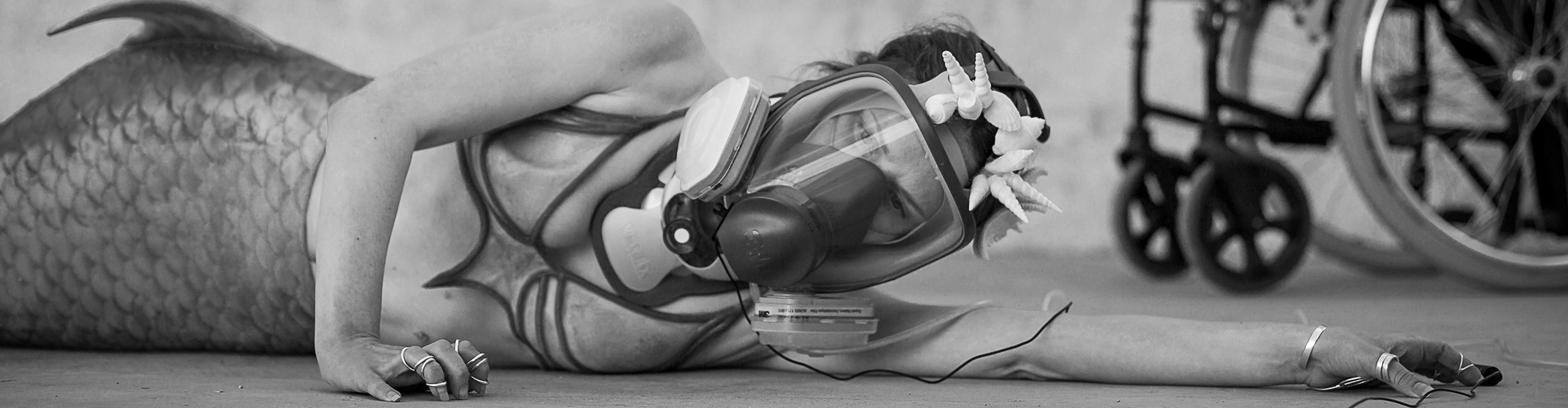 A mermaid lies on her side with one arm outstretched along the ground above of her head. She wears a gas mask, a neck brace and shells in her hair. The wheels of a wheelchair are in the background.