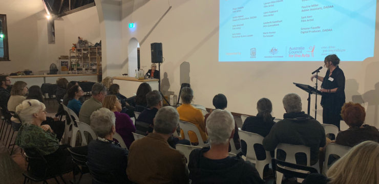 About 30 people sit on chairs in a hall. They look at the wall onto which a screen is projected with the Arts Access Australia logo. A woman in her 50s (Meagan Shand) speaks into a microphone.