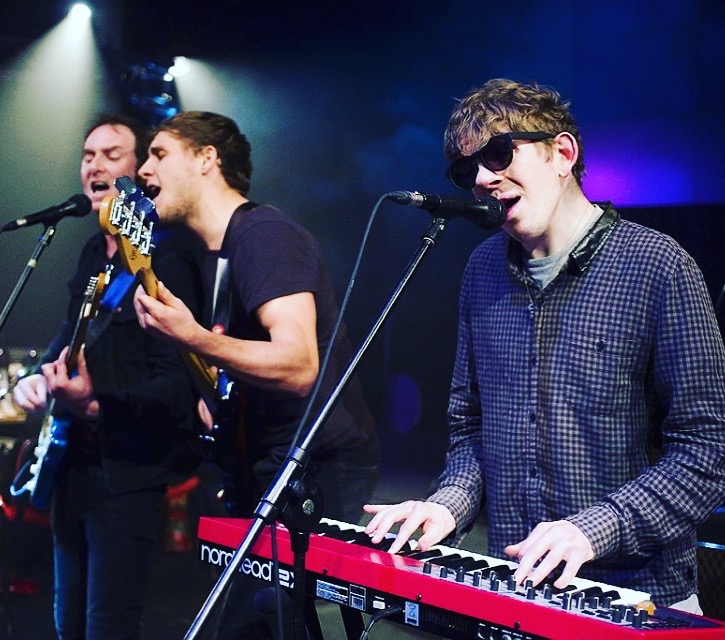 Three musicians perform on stage - two on guitars singing into the same mike, and one on keyboards wearing dark glasses and singing into a mike.