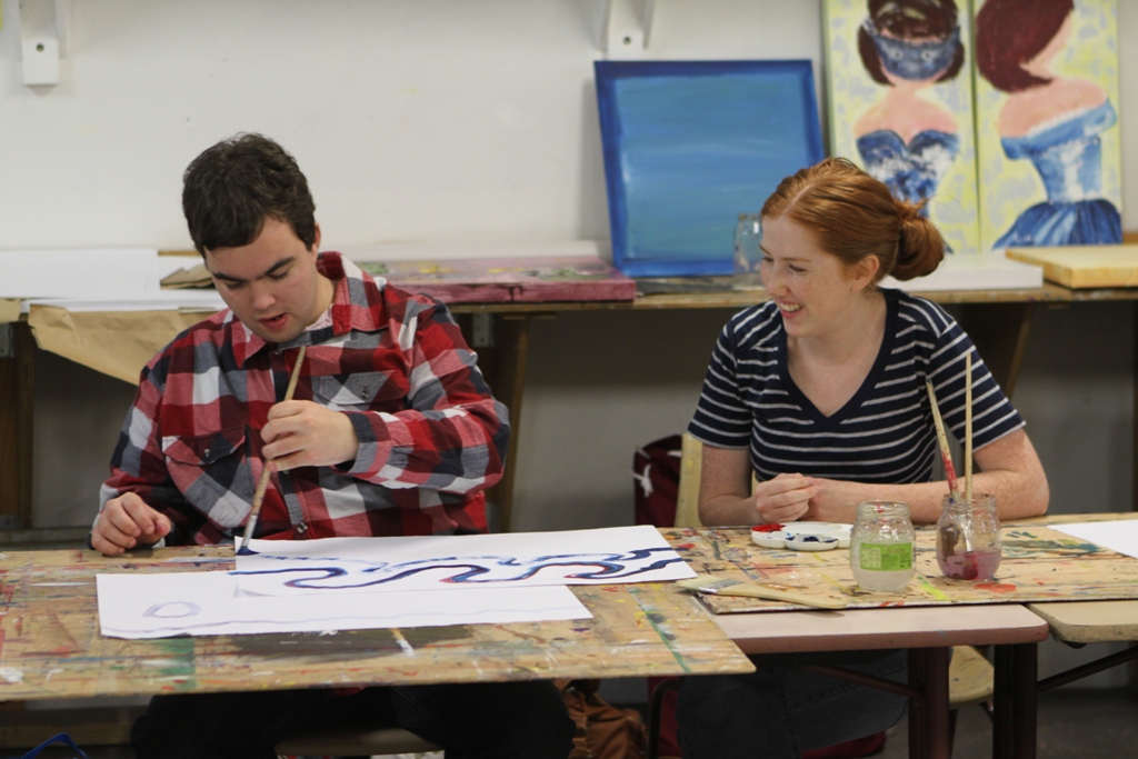 A man in his twenties paints at a desk alongside a woman with red hair, also in her twenties. She is looking over at him and smiling.
