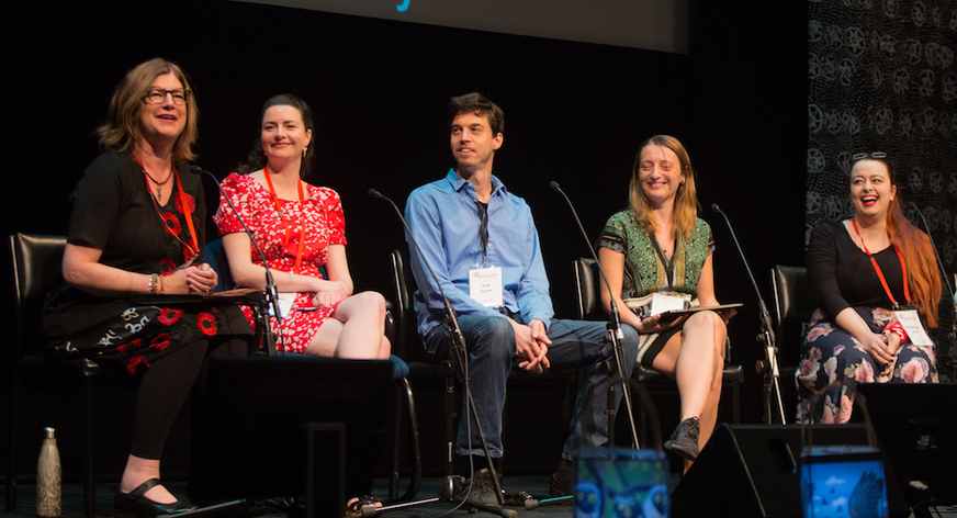 Five people sit on chairs on a stage. On the far left sits a woman in her 50s who is talking. The other panel members (three females and one male) look toward the woman and smile.