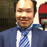 A young man of Asian heritage smiles at the camera. He wears a blue suit with a blue and white striped tie.