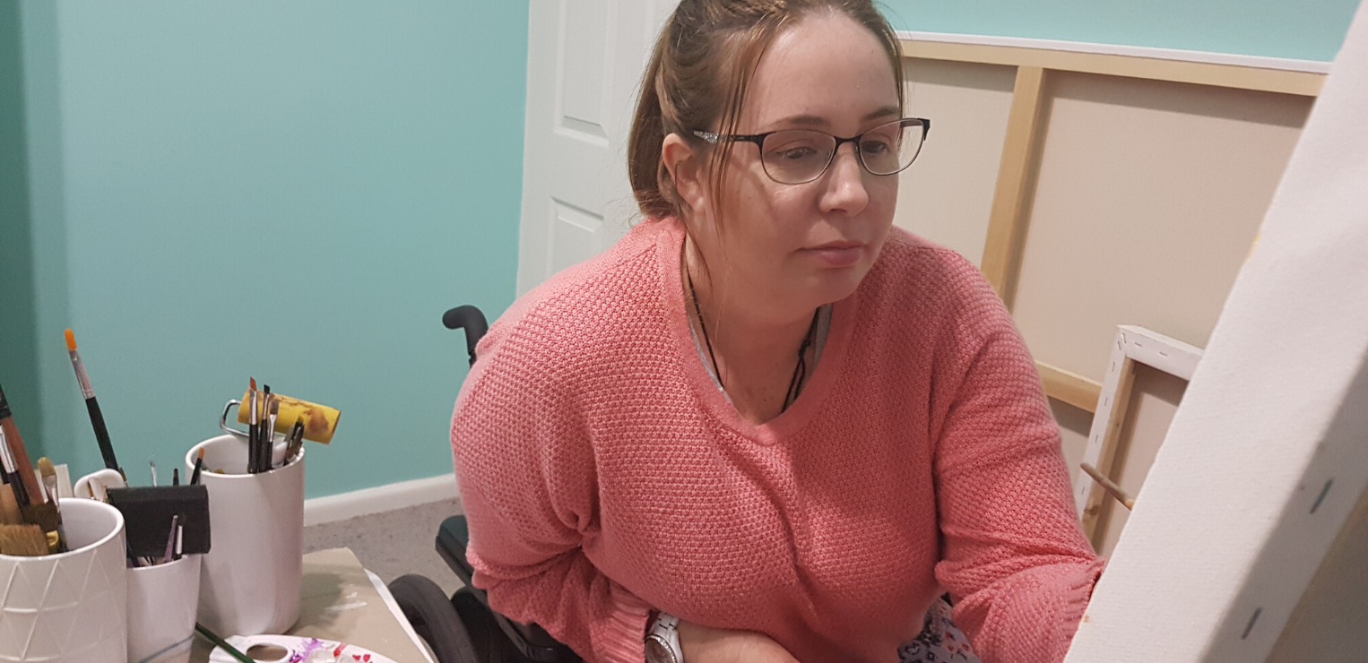 A woman aged in her late 30's wearing glasses, in a pink jumper sitting in her wheelchair. Her hair is tied back and she is looking at a canvas and holding her paintbrush. There are paints to the side of her and she looks deep in concentration.