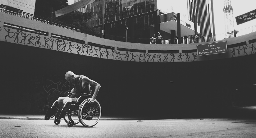 A black and white photo of a man using a manual wheelchair in an urban environment.