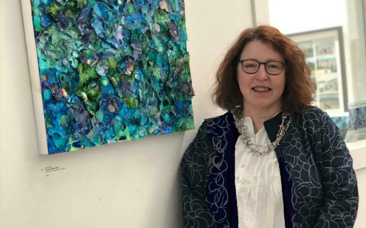 A white skinned woman in her 50's, red/brown shoulder length curly hair, glasses, smiling at the camera while standing in front of a green, blue artwork.