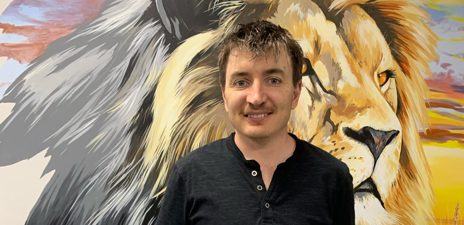 A white skinned male with short brown hair and a moustache. He stands in front of a painting of a lion. He is wearing a black shirt and jeans.