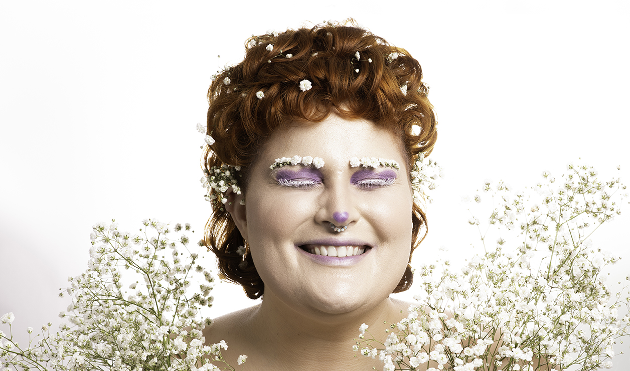 A white person with ginger curly hair and purple makeup adorned in a halo of babies breath on their bare chest. Their hair also has pieces of babies breath decorated within it, with a small branch of it tucked behind their ear. Their eyebrows have also been covered in babies breath. Their eyes are closed and are smiling. The background is white.
