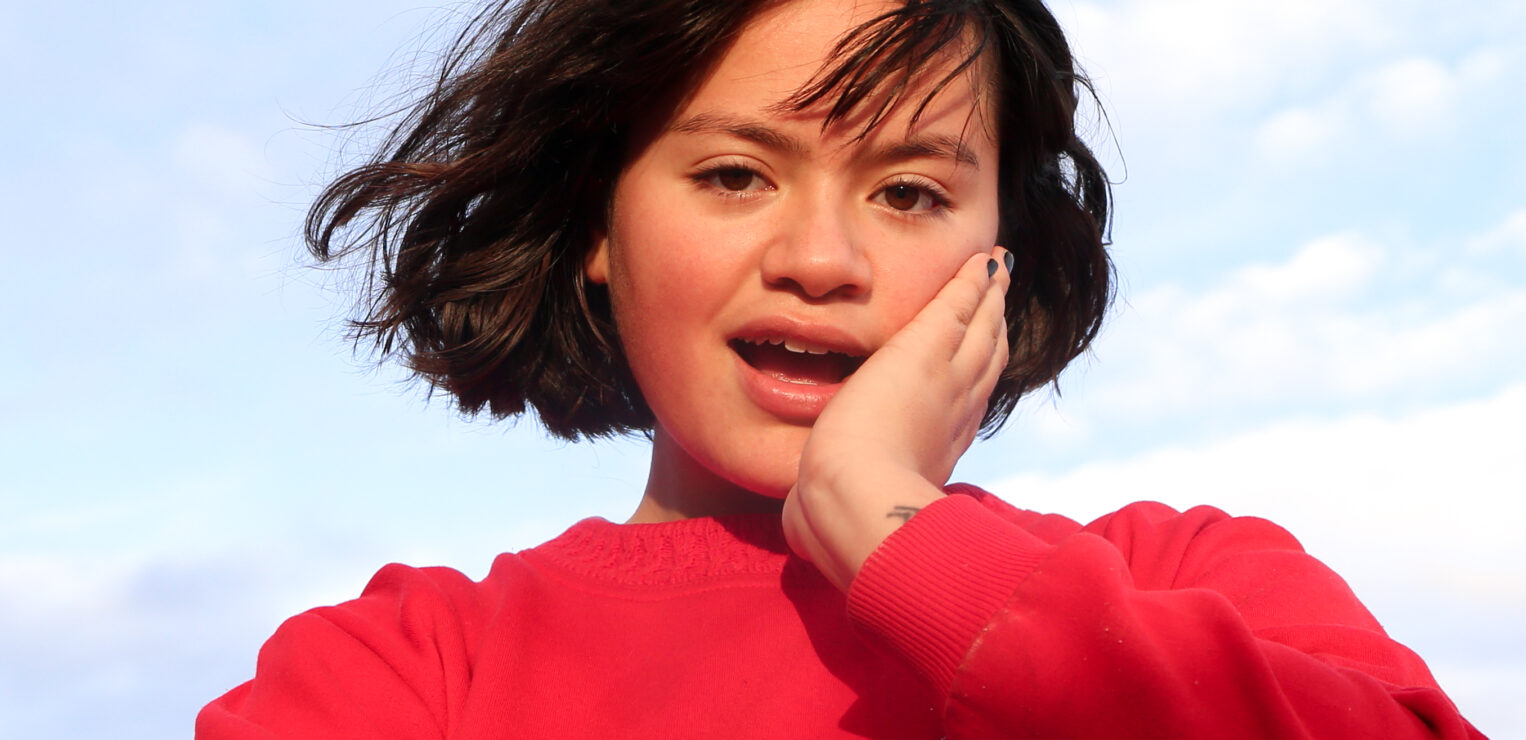 A light brown skinned woman in her 20s with dark wavy chin length hair and a fringe. She is wearing a bright red jumper and is standing in front of blue sky. She has one arm crossed and the other cupping her face. 
