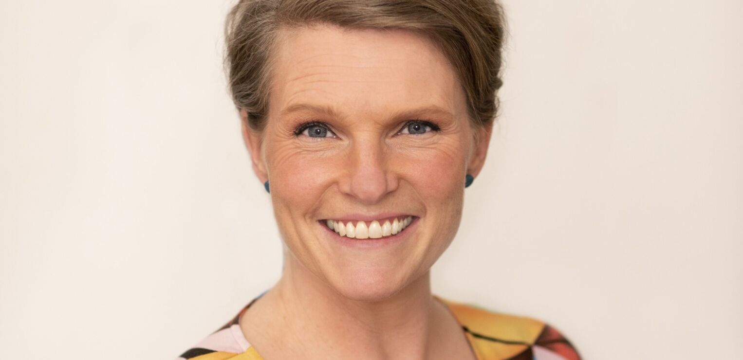 A woman with white skin and short brown blonde hair smiles. She has blue earrings and a multicoloured top on.