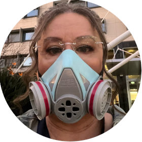 A woman with glasses and a blue and pink respirator looks into tghe camera